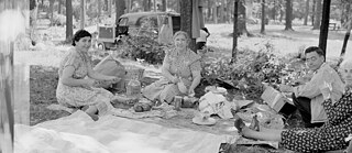 Ein Schwarz-Weiß-Foto einer Gruppe von Personen, die in einem Waldgebiet ein Picknick machen, auf einer Decke sitzen und Essen um sich herum verteilt haben. Im Hintergrund steht ein Fahrzeug.