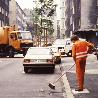 Foto di altri tempi che mostra la raccolta delle immondizie a Dortmund. Si vede un operatore ecologico in uniforme arancione intento a spazzare la strada, mentre sullo sfondo si nota il camion della spazzatura.