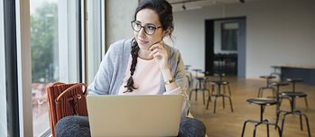 Eine Frau sitzt mit ihrem Laptop auf einem Podest am Fenster. Sie sitzt im Schneidersitz und lächelt in die Kamera.