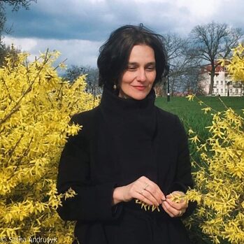 Foto af Katja Petrowskaja, der står mellem to forsythia-buske og smilende ser nedad.