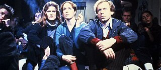 Plusieurs jeunes sont assis par terre dans une salle obscure, ils fument et regardent un film projeté par un projecteur.