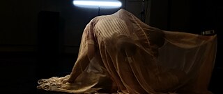 Two dancers are covered in a white fabric. The performance takes place in a dark envorinoment, lit by a fluorescent lamp.