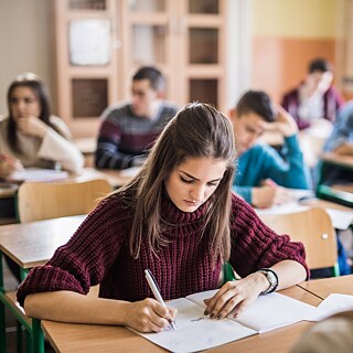 Schüler schreiben einen Test im Klassenraum. Der Fokus liegt auf einer Schülerin im Vordergrund.