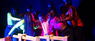 CONTEXTOS | MEMORIAS | Eight dancers behind Stool-wall. 8 dancers from Memorias Danza Teatro are performing the piece KZ El Religar de las Almas. In the photograph, the dancers are together, but the stool has been placed between them, marking a space of separation or resistance. The bodies appear to be in a moment of transition, as if preparing for a change or confrontation.  Place: Sala de teatro Reculá del Ovejo.