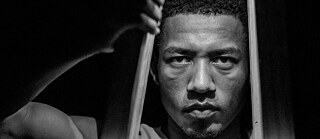 CONTEXTOS | MEMORIAS | One Person with stool:  The dancer Luis David Ochoa from Memorias Danza Teatro Company is performing the piece KZ El Religar de las Almas. | In this photograph, a single dancer is seen standing in front of the stool. The posture is strong, with the legs open and the hands on the sides of the stool, almost as if he/she were embracing it or claiming his/her space. The lighting highlights the figure of the dancer, leaving the background in shadow, which generates an atmosphere of introspection and resistance. The proximity to the stool emphasizes the symbolic relationship between the body and the object. | Place: Sala de teatro Reculá del Ovejo. 