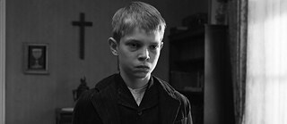 Das Schwarz-Weiß-Bild aus dem Film „Das weiße Band“ zeigt eine Nahaufnahme des Gesichts eines Jungen. Sein Gesichtsausdruck ist hart und traurig zugleich. Im Hintergrund ist ein großes Kruzifix zu sehen