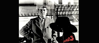 Black and white image of a man looking into a shop window with a display of hats
