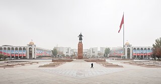  Ein Frau geht durch einen Platz in Bischkek mit einer Statue in der Mitte, umgeben von Bäumen und Gebäuden.