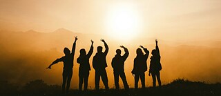 Eine Gruppe von Menschen als Silhouette vor einem Sonnenuntergang, die ihre Arme in Freude und Gemeinschaft in die Höhe streckt.