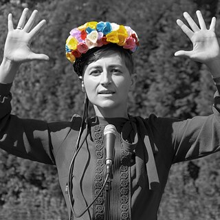 Portrait of a woman. Her mouth is slightly open, and she is looking into the camera. A microphone is visible in front of her. The image is black and white, with only her headpiece showing a colorful flower crown.