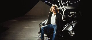 Un acteur sur scène est assis sur une voiture noire dont des branches blanches sortent du capot.