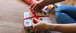 A person sits on the floor and tightens the red ribbon of a present. There are rolls of wrapping paper in the background. 