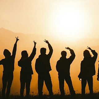 Eine Gruppe von Menschen als Silhouette vor einem Sonnenuntergang, die ihre Arme in Freude und Gemeinschaft in die Höhe streckt.