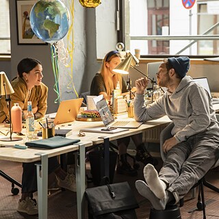 Tim (Frederick Lau) und eine Kollegin unterhalten sich, während sie an einem Tisch im Büro sitzen.