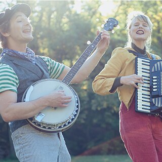 ein Mann spielt auf einer Art Gitarre und singt, neben ihm eine Frau, die Akkordeon spielt und mitsingt, die beiden stehen in der Natur 