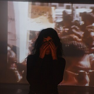 A person stands with hands over face in a dimly lit room, with a projected image of a crowded protest scene in the background.