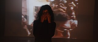 A person stands with hands over face in a dimly lit room, with a projected image of a crowded protest scene in the background.