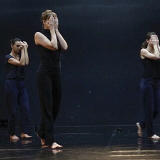  Quatre danseuses traversent la scène avec recueillement et cachent leurs visages avec leurs mains. L’arrière-plan est noir, seulement la scène, qui est en noir couleur aussi est illuminée.
