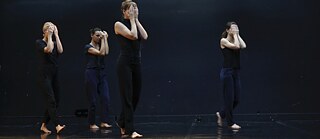  Quatre danseuses traversent la scène avec recueillement et cachent leurs visages avec leurs mains. L’arrière-plan est noir, seulement la scène, qui est en noir couleur aussi est illuminée.