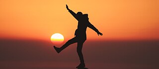 Silhouette of a person standing on a rock with one leg raised and arms outstretched against a sunset. © © Unsplash Deutsch verbessern mit den richtigen Tools