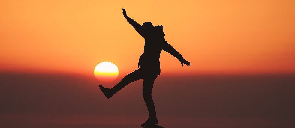 Silhouette of a person standing on a rock with one leg raised and arms outstretched against a sunset.