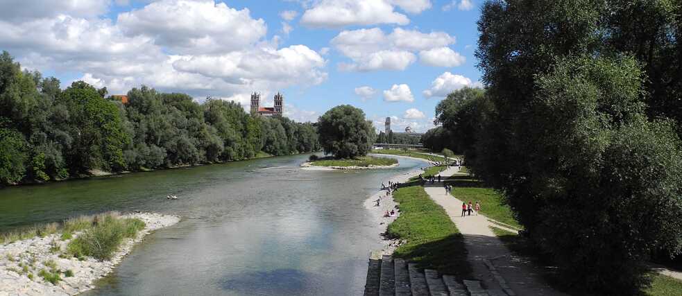 Eine Flusslandschaft mit Kiesstrand, an dem sich Menschen aufhalten. Im Bildhintergrund eine Kirche.   