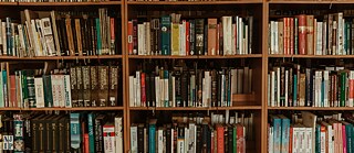 A cozy library interior with tall wooden bookshelves filled with books of various sizes and colors. Comfortable reading chairs and tables are arranged in the center, with soft natural light streaming through large windows. The atmosphere is quiet and inviting, perfect for reading and studying