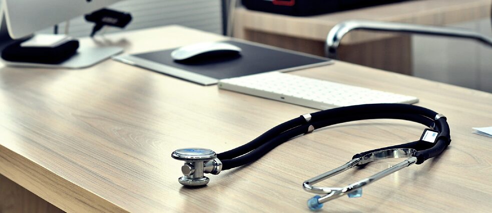 A wooden desk with a stethoscope placed on it, alongside a computer setup including a monitor, keyboard, and mouse. A printer is visible in the background, suggesting a medical office environment