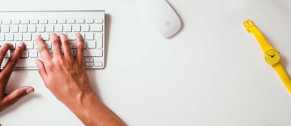 A person typing on a white keyboard, with a yellow wristwatch and a white computer mouse on the desk.