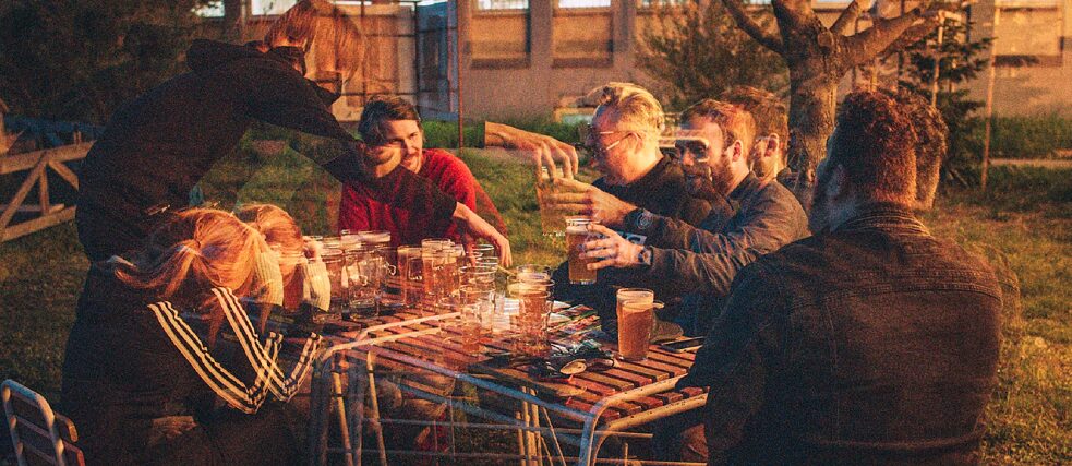 A group of people sitting outdoors at a table, enjoying drinks together. The image has a double-exposure effect.