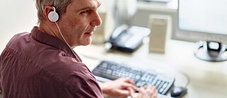 Ein Mann sitzt am Schreibtisch und liest an einer Braillezeile. Er trägt einen Ohrhörer.