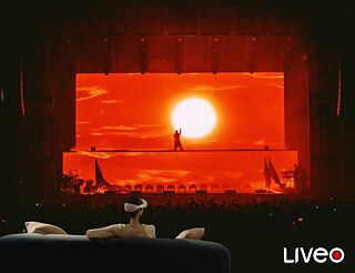 Im Vordergrund sitzt jemand auf einem Sofa. Die Person hat eine VR-Brille auf. Im Hintergrund sieht man die Projektion einer Show.