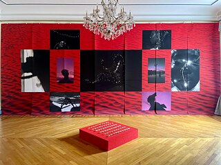 Zu sehen ist ein Wand in einem Ausstellungsraum, die mit großflächigen Foto-Arbeiten bedeckt ist. Davor ist ein Kunstwerk auf einem roten Sockel ausgestellt. 