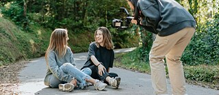 A man films two young women sitting on the ground, capturing their joyful expressions in a relaxed outdoor scene.