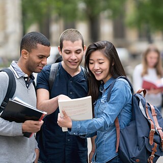 Drei Studenten unterhalten sich in einem Park. Sie betrachten gemeinsam ein Buch und reden darüber. Sie tragen Rucksäcke und Unterlagen.
