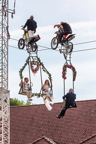A wedding couple on a swing suspended from a tightrope and pulled by two men on motorbikes. 