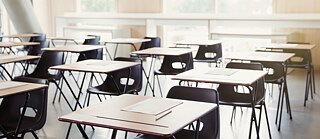 Ein leerer Klassenraum mit quadratischen Tischen in Reih und Glied. Auf jedem Tisch liegen säuberlich angeordnet ein paar Blätter Papier