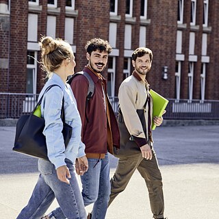 Fero, Noah und Tatjana auf dem Weg zum Goethe-Institut Hamburg. Die Gruppe geht über den Vorplatz. Sie unterhalten sich. Noah hält eine Mappe der Goethe-Boutique in der Hand Tatjana hat eine Trinkflasche in der Tasche.