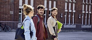 Fero, Noah und Tatjana auf dem Weg zum Goethe-Institut Hamburg. Die Gruppe geht über den Vorplatz. Sie unterhalten sich. Noah hält eine Mappe der Goethe-Boutique in der Hand Tatjana hat eine Trinkflasche in der Tasche.
