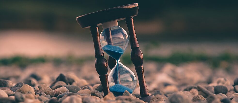 A close-up photograph of an hourglass with blue sand, resting on small stones. The hourglass is tilted, with sand trickling down, symbolizing the passage of time. The background is blurred, creating a warm and serene atmosphere.