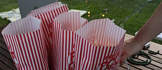 Several red-and-white striped popcorn bags placed on a wooden table, with a person's hand reaching out. The background features green grass, fairy lights, and a cozy outdoor setting, suggesting a movie night or outdoor gathering. © © Unsplash Movie Night Snacks