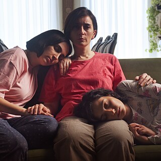 Im Vordergrund ist die Tochter des Anwalts zu sehen. Ihr Blick ist ernst. Dahinter unscharf ihre Mutter und Schwester, 