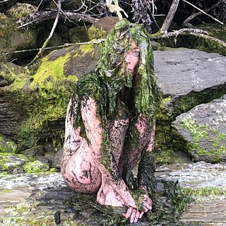 Eine Frau sitzt auf Felsen. Ihr Kopf ist von nassen Pflanzen bedeckt.