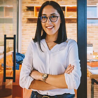 Eine Geschäftsfrau steht selbstbewusst mit verschränkten Armen in einem modernen Büro und blickt in die Kamera.