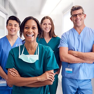 Pflgekräfte in blauen und grünen Uniformen stehen auf dem Krankenhausflur und lachen in die Kamera.