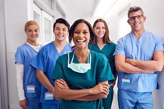 Pflgekräfte in blauen und grünen Uniformen stehen auf dem Krankenhausflur und lachen in die Kamera.