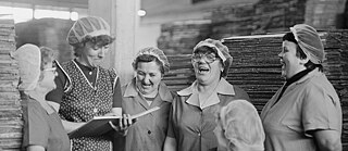 Ein Schwarz-Weißfoto aus einer Fabrik in der DDR mit 6 Frauen in Arbeitskleidung, die zusammen stehen und lachen
