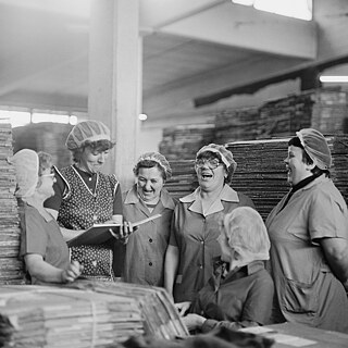 Ein Schwarz-Weißfoto aus einer Fabrik in der DDR mit 6 Frauen in Arbeitskleidung, die zusammen stehen und lachen