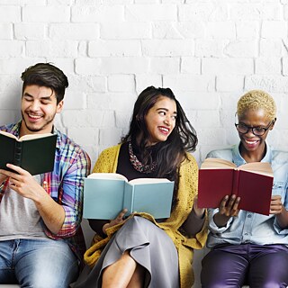Mehrere junge Studenten sitzen nebeneinander auf Stühlen und lesen in Büchern. Sie lachen herzlich.