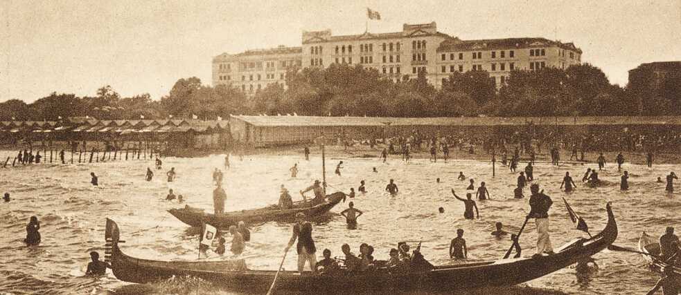 Photo postcard “Grandhôtel des Bains” – Venice, Lido; in the background a large building, in front of it a body of water with numerous people bathing and two Venetian gondolas